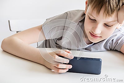 Teen boy sits at white table and uses smartphone Stock Photo