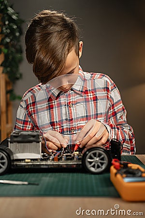 Teen boy sitting at table and soldering remote controlled car. Caucasian child fixing favorite toy by himself at home. Stock Photo