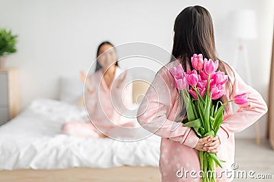 Teen girl hold bouquet of tulips behind back and congratulates asian millennial woman in bedroom interior Stock Photo