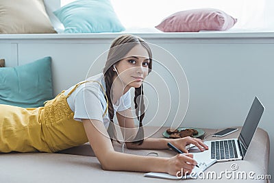 teen girl lying on sofa with laptop textbook smartphone earphones Stock Photo