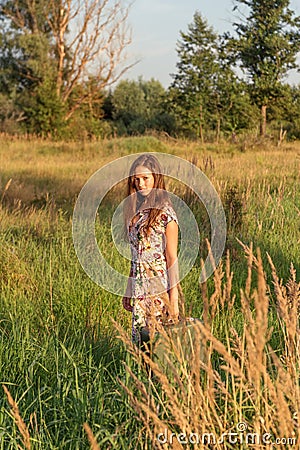 A young girl in a retro vintage dress with flowing hair is standing in a tall grass in a sunlight afterglow with a suitcase Stock Photo