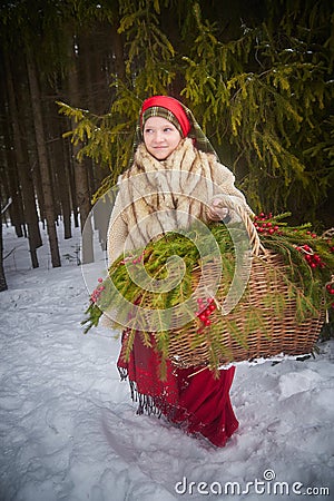 Teen girl in thick coat and a red sash with basket of fir branches and berries in cold winter day in forest. Medieval Stock Photo
