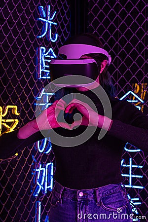 Teen girl using vr helmet shows heart gesture with hands on futuristic neon colorful background. Virtual love concept Stock Photo