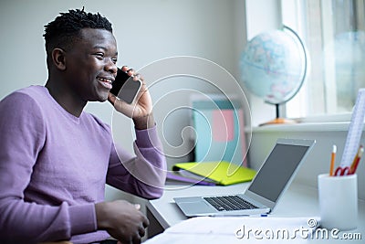 Teenage Boy Sitting At Desk Doing Homework Assignment On Laptop Talking On Mobile Phone Stock Photo