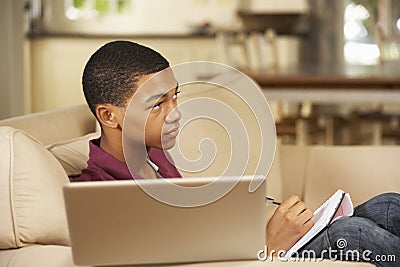 Teenage Boy Sitting On Sofa At Home Doing Homework Using Laptop Computer Whilst Watching TV Stock Photo