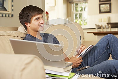 Teenage Boy Sitting On Sofa At Home Doing Homework Using Laptop Computer Whilst Watching TV Stock Photo