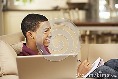 Teenage Boy Sitting On Sofa At Home Doing Homework Using Laptop Computer Whilst Watching TV Stock Photo