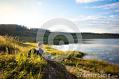 A teenage boy travels on foot. A young man sits on a stone on the shore of a lake in a beautiful place. Hiking Stock Photo