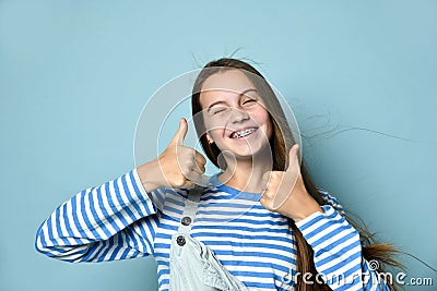 Teenage female in jeans overall, striped sweatshirt. She smiling, showing thumbs up, winking, posing on blue background. Close up Stock Photo