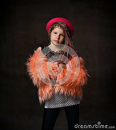 Teenage girl with african pigtails, in red hat, orange faux fur coat. Propping her chin by forefinger, posing on dark background Stock Photo
