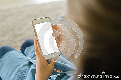 Teenage girl in a blue dress sitting on an armchair while holding a smart phone with a blank screen in the hands. Young girl using Stock Photo