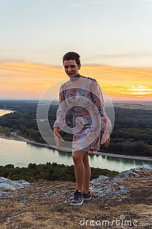 teenage girl in dress, with short-cropped hair, on top cliff, on river bank, at sunset Stock Photo
