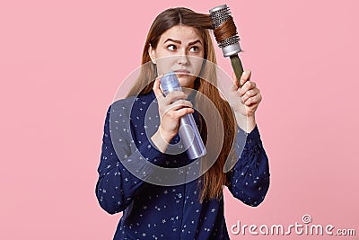 Teenage girl hurries for date, tries to make hairstyle, combs hair with hairbrush, uses hairspray, dressed in shirt, poses over Stock Photo