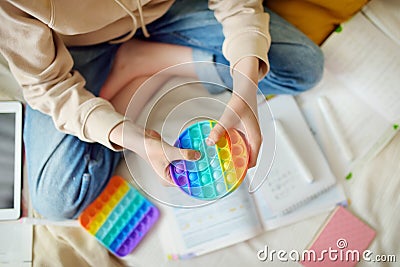 Teenage girl playing with rainbow pop-it fidget toy while studying at home. Teen kid with trendy stress and anxiety relief Stock Photo