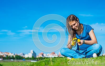 Teenager brunette girl with long hair sit on the grass and wreathes a wreath of yellow dandelion flowers on sky background with co Stock Photo
