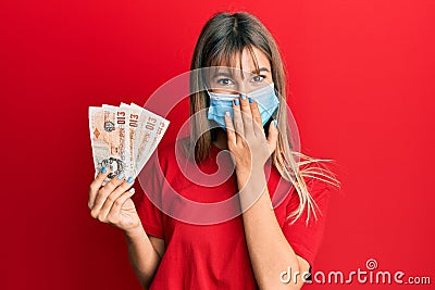 Teenager caucasian girl wearing medical mask holding 10 united kingdom pounds banknotes covering mouth with hand, shocked and Editorial Stock Photo
