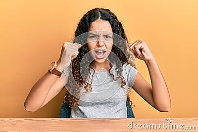 Teenager hispanic girl wearing casual clothes sitting on the table angry and mad raising fists frustrated and furious while Stock Photo