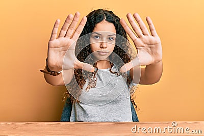 Teenager hispanic girl wearing casual clothes sitting on the table doing frame using hands palms and fingers, camera perspective Stock Photo