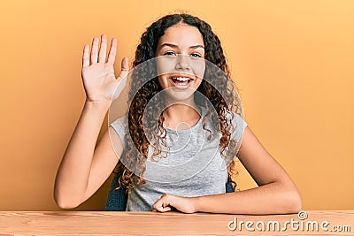 Teenager hispanic girl wearing casual clothes sitting on the table waiving saying hello happy and smiling, friendly welcome Stock Photo