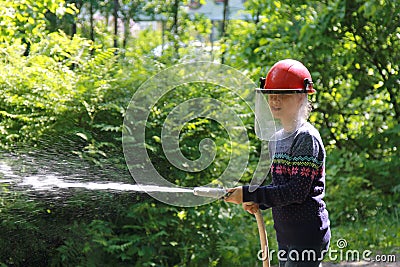 Teenager learning the firefighter profession. The girl in fire helmet pours water from the hose Stock Photo