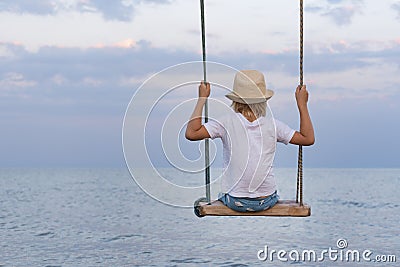 Teenager in straw hat on sea background. Back view. Rope swing over water. Calm and relaxing Stock Photo