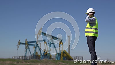 Teknikern på telefonen nära olje- pumpar som anmäler de tillgängliga extraktioninstallationerna för rå oljor, levererar funktions lager videofilmer Teknikern på telefonen nära olje- pumpar som anmäler de tillgängliga extraktioninstallationerna för rå oljor, levererar funktions lager videofilmer