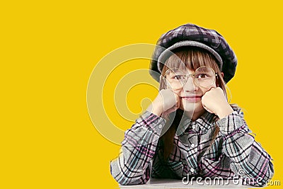 A ten-year-old girl in a plaid hat and a plaid shirt props up her chin with her hand, wearing glasses, close-up, yellow background Stock Photo