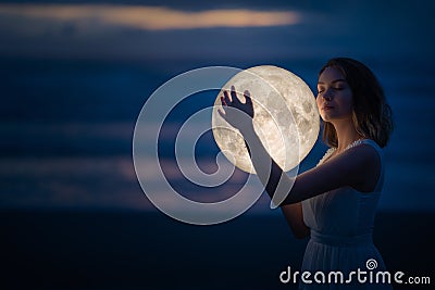 Tender image of a girl. Female magic. Beautiful attractive girl on a night beach with sand hugs the moon, art photo. On a dark Stock Photo