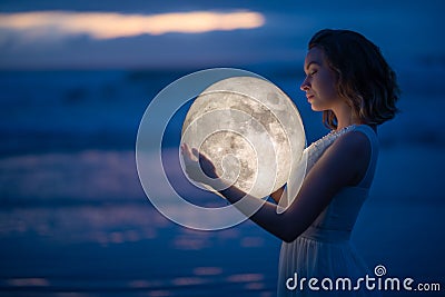 Tender image of a girl. Female magic. Beautiful attractive girl on a night beach with sand hugs the moon, art photo. On a dark Stock Photo