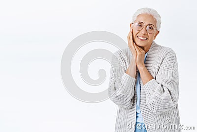 Tender lovely senior woman with grey combed hair in prescription glasses, cozy cardigan, touching cheek and smiling Stock Photo