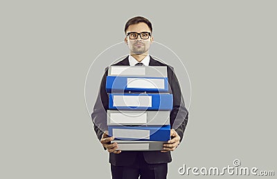 Tense male office worker with stressed expression holding stack of heavy folders on gray background. Stock Photo