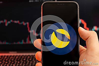 Terra LUNA cryptocurrency logo on the screen of smartphone in mans hand with downtrend on the chart on a red light Editorial Stock Photo