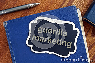 Text sign showing guerilla marketing. The text is written on a small wooden blackboard. The book, pen, wooden background are on Stock Photo