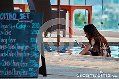 THAILAND, PHUKET, MARCH 19, 2018 - Girl uses her smartphone in pool by the bar. Rear view. Concept dependence on social networks. Editorial Stock Photo