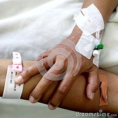 There is a middle-aged woman sore hand with a close-up infusion set and a wristband on a white hospital bed sheet. Stock Photo