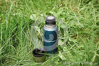 Thermos and a mug with a hot drink standing on the grass outdoors, top view. Steam rising from a steel cup Stock Photo