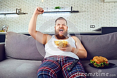 Thick funny man in pajamas eating a burger sitting on the couch. Stock Photo