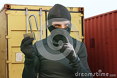 Thief at work - criminal man in black covered with balaclava mask holding unlocked padlock at shipping area break in storage Stock Photo