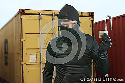 Thief at work - criminal man in black covered with balaclava mask holding unlocked padlock at shipping area break in storage Stock Photo