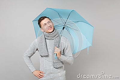 Thinkful young man in gray sweater, scarf looking up, holding blue umbrella isolated on grey background. Healthy fashion Stock Photo