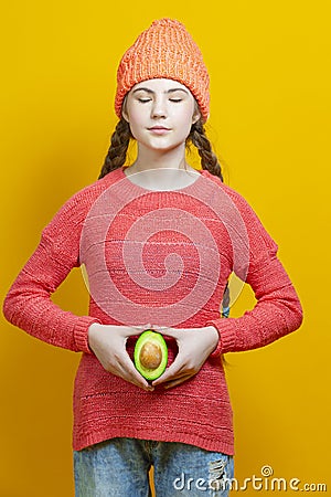 Female Fertile Concepts. Thinking Caucasian Girl In Coral Knitted Clothing Posing With Split Avocado Fruit In Front of Belly as a Stock Photo