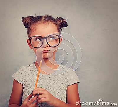 Thinking grimacing kid girl in glasses looking and holding pencil in hand on blue background with empty copy space. Toned vintage Stock Photo