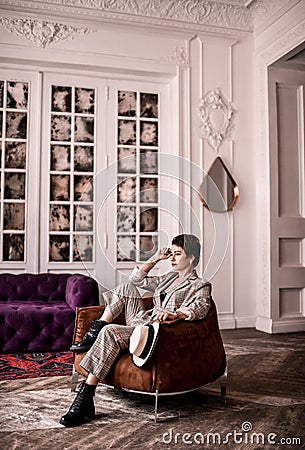 Thinking short-haired brunette woman in elegant business plaid pantsuit with rolled up sleeves sits in armchair in room Stock Photo