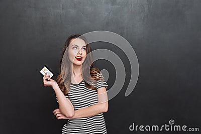 Thinking young lady standing over grey wall holding debit card Stock Photo