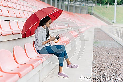 Thoughtful afro-american teenager under the red umbrella. She is chatting via the mobile phone. Side view. Stock Photo