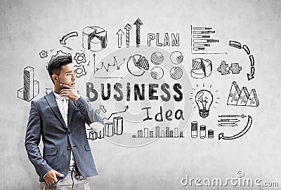 Thoughtful Asian businessman is standing near a concrete wall with black business idea sketch. Stock Photo