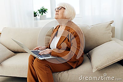 a thoughtful, enthusiastic elderly woman in a stylish brown suit sits on a beige sofa with a laptop on her lap and looks Stock Photo