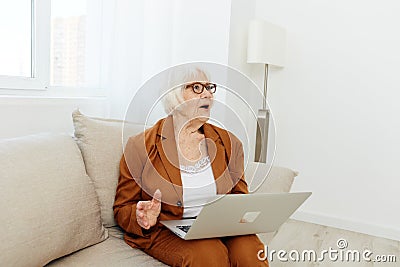 a thoughtful, enthusiastic elderly woman in a stylish brown suit sits on a beige sofa with a laptop on her lap and looks Stock Photo