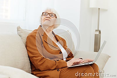 a thoughtful, enthusiastic elderly woman in a stylish brown suit sits on a beige sofa with a laptop on her lap and looks Stock Photo