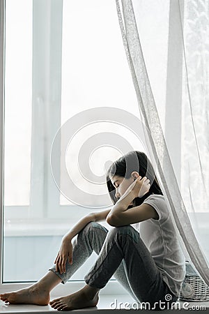 A thoughtful European girl of 8 years old sitting on a window sill. Stock Photo